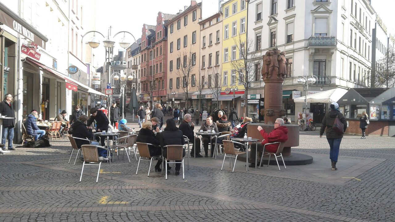 An der Kreuzung von Berger- und Wiesenstraße ist ein dreieckiger Platz mit Denkmal im Zentrum. Menschen sitzen in dicken Jacken draußen an Tischen, die Sonne scheint fahl und die Bäume sind noch kahl.