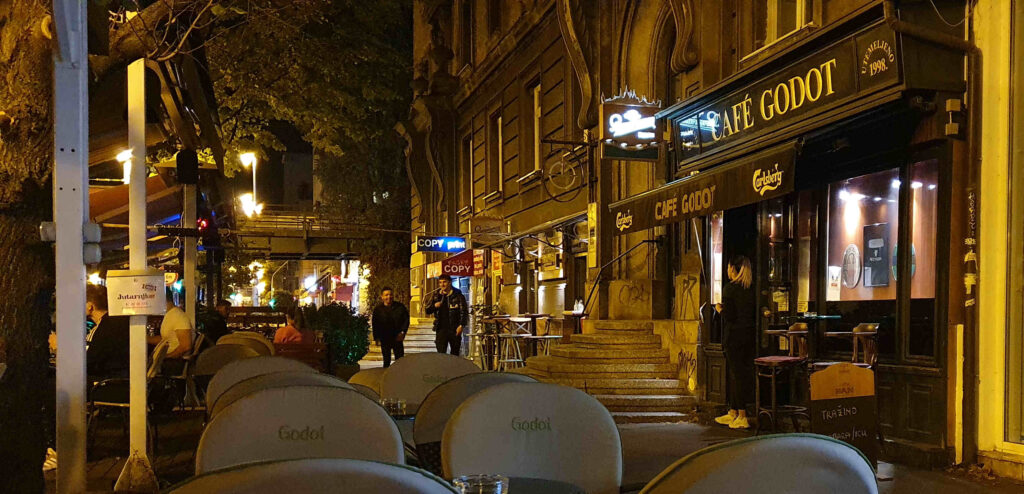 Nächtlicher Bürgersteig mit Passanten, im Vordergrund Cafétische, rechts das Lokal Café Godot, Gründerzeithäuser und im Hintergrund eine Bahnüberführung