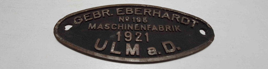 Ovales Produktionsschild einer Dampflokomotive mit Gebrauchsspuren aus Gusseisen, der Text lautet: "Gebr. Eberhardt / No. 195 / Maschinenfabrik / 1921 / Ulm a. D."