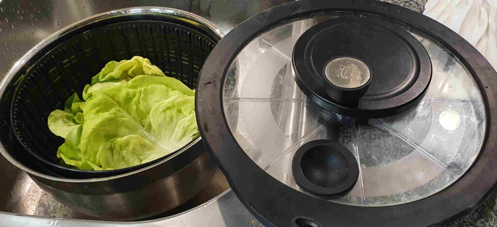 Links gelbgrüne Kopfsalatblätter mit weißer Mittelrippe in einem schwarzen Plastikkorb, der wiederum in einer Edelstahlschüssel steht, rechts der Deckel der Salatschleuder mit Kurbel